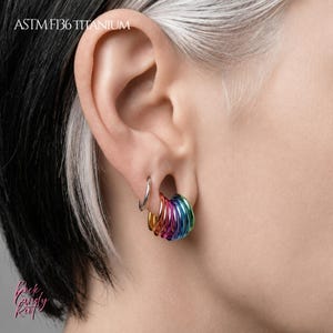 May include: A set of ASTM F136 titanium ear rings featuring a single plain silver-toned hoop and a cluster of multiple vibrant, rainbow-colored anodized hoops. The jewelry is displayed in a stretched earlobe, showcasing a metallic finish.