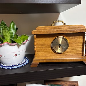 May include: A wooden mantel clock with a gold-colored clock face and a brass handle. The clock is on a dark shelf next to a white ceramic pot with a green plant. The pot has a pink rim and red floral designs.