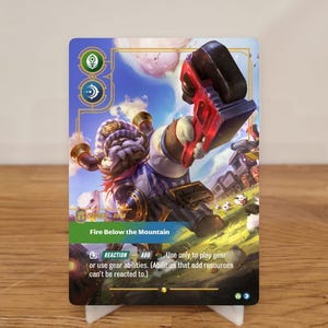 Könnte beinhalten: Sammelkarte mit einem Fantasy-Charakter, der ein rot-schwarzes Gerät hält. Die Karte enthält den Text "Fire Below the Mountain" und Spielanweisungen. Der Hintergrund zeigt eine Landschaft mit einem Zug.