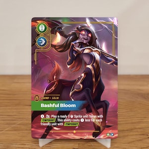 May include: A trading card featuring a fantasy character, Bashful Bloom, with a centaur-like lower body and a human upper body. The card has a green banner with the name and text describing the character's abilities. The card is propped up on a white stand.