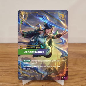 Könnte beinhalten: Eine Sammelkarte mit einer Frau in einem fließenden teal- und weißen Outfit mit blauen Energieeffekten. Die Karte trägt die Aufschrift "Defiant Dance" und enthält Spielanweisungen. Die Karte steht auf einem weißen Ständer.