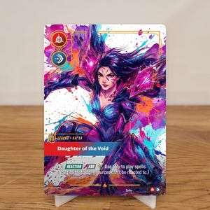 May include: A trading card featuring a female character with long dark hair and a blue and purple outfit. The card reads "LEGEND - KAI'SA" and "Daughter of the Void." The card is on a white stand.