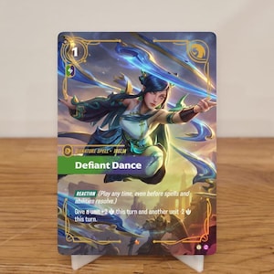 Op de afbeelding: Een kaart met een vrouw in blauw en groen, met de tekst "Defiant Dance". De kaart heeft een blauw en goud kleurenschema, met het nummer "1" bovenaan. De kaart staat op een witte standaard.