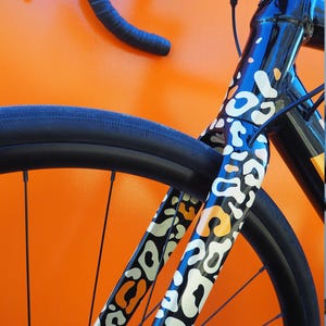 Puede incluir: Primer plano de una bicicleta negra con un llamativo diseño de estampado de leopardo en blanco, naranja y negro en el cuadro. La bicicleta tiene neumáticos y manillar negros, sobre un fondo naranja.
