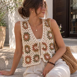 Pode incluir: Um colete de crochê sem mangas com padrão de quadrados granny em creme, verde sálvia e rosa antigo. O colete tem um decote em V profundo com fecho de laço central e bainha curta, usado sobre calças claras e combinado com uma bolsa.