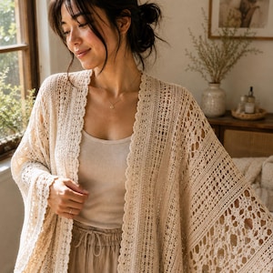 Può includere: Un cardigan all'uncinetto beige aperto sul davanti con un intricato motivo in pizzo e bordi smerlati, indossato sopra una canotta e pantaloni con coulisse in tinta, completato da una delicata catenina d'oro con un piccolo ciondolo.