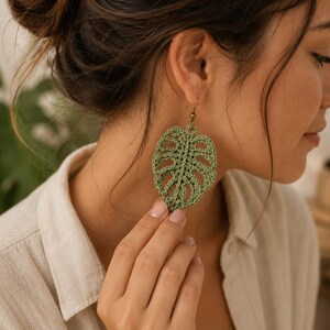 May include: A sage green crochet earring shaped like a monstera leaf with intricate openwork detailing. The earring features a bronze-toned metal hook for attachment, showcasing a detailed fiber art design with a distinct leaf vein pattern.