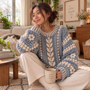 May include: A long-sleeved crochet sweater in light blue and cream, featuring a central vertical leaf pattern, horizontal rows of cream bobbles, and floral motifs on the sleeves. The sweater has a ribbed crew neckline and cuffs.