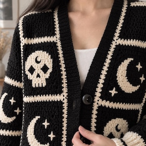 Puede incluir: Un cárdigan de ganchillo en color negro y crema con un patrón cuadrado repetitivo de calaveras, lunas crecientes y estrellas. La prenda tiene un cuello en V, una tapeta frontal acanalada y grandes botones negros en el centro.