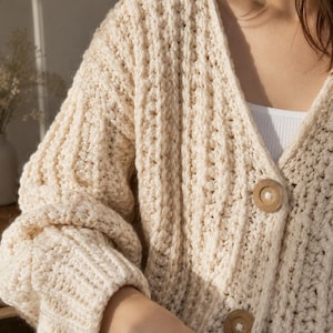 Könnte beinhalten: Eine cremefarbene, grob gestrickte Strickjacke mit tiefem V-Ausschnitt, langen Ärmeln mit gerippten Bündchen und großen, runden, hellbraunen Holzknöpfen auf der Vorderseite. Das Material ist dick und durchgehend vertikal gerippt.