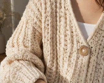 Puede incluir: Un cárdigan de punto grueso en color crema con un escote en V profundo, mangas largas con puños acanalados y grandes botones redondos de madera de color marrón claro en la parte delantera. La textura es gruesa y acanalada verticalmente.