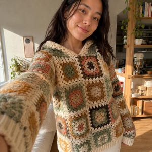 Puede incluir: Un suéter con capucha y mangas largas confeccionado con un mosaico de cuadrados de ganchillo en tonos apagados de crema, verde salvia, rosa viejo y amarillo mostaza, con un dobladillo y puños acanalados de color crema.