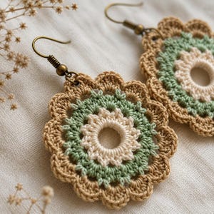 May include: A pair of round crochet earrings featuring a layered floral design. The center is cream, surrounded by a sage green ring, and finished with a tan scalloped edge. Each earring hangs from a bronze-toned metal hook for pierced ears.