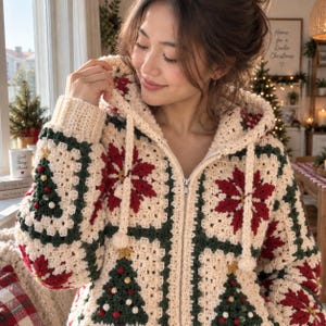 Op de afbeelding: Een crèmekleurige gehaakte hoodie met rits, voorzien van een granny square patroon met afwisselend rode kerststerren en groene kerstbomen. De hoodie heeft een capuchon met trekkoord met pompons en geribbelde boorden aan de mouwen.