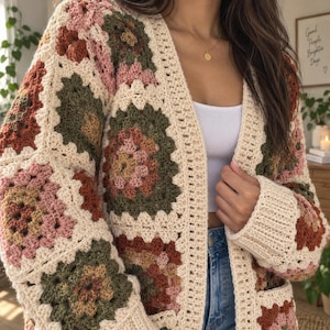 Puede incluir: Un cárdigan de ganchillo de manga larga confeccionado con cuadrados de la abuela multicolores en tonos crema, verde oliva, rosa empolvado y terracota. La prenda presenta un borde acanalado de color crema en la abertura frontal y los puños.
