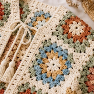 May include: A crochet cardigan constructed from multi-coloured granny squares in shades of cream, sage green, dusty blue, and terracotta. The garment features a front tie closure with two long cream-coloured yarn tassels hanging from the neckline.