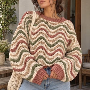 May include: A long-sleeve crochet sweater featuring a wavy, multi-colored pattern in shades of dusty rose, sage green, and cream. The garment has a ribbed crew neckline and ribbed cuffs, paired with blue denim jeans and a cream crochet bag.