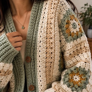 Könnte beinhalten: Eine Häkelstrickjacke aus salbeigrünem, cremefarbenem und senfgelbem Garn. Das Kleidungsstück zeigt eine Kombination aus einfarbigen Rippenstrickbahnen und komplexen, blumenartigen Granny-Square-Motiven, abgerundet durch zwei braune, runde Knöpfe.