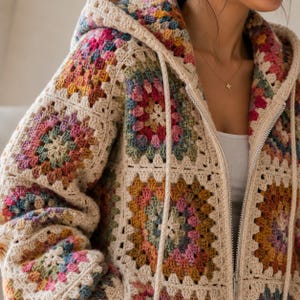 May include: A zip-up hoodie constructed from crocheted granny squares in a palette of cream, mustard yellow, dusty pink, sage green, and muted blue. The garment features a hood with matching cream-colored drawstrings and a silver metal zipper.