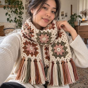 Op de afbeelding: Een gehaakte sjaal met een herhalend granny square patroon in tinten crème, oudroze, olijfgroen en terracotta. De sjaal is afgewerkt met lange, dikke garenkwasten aan beide uiteinden, afgewisseld in hetzelfde kleurenpalet van crème, roze, groen en bruin.