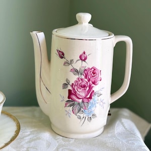 Könnte beinhalten: Cremefarbene Vintage-Kaffeekanne mit Deckel, Griff und Ausguss. Die Kanne ist mit rosa Rosen, blauen Blumen und grünen Blättern verziert. Der Rand und der Ausguss sind mit Gold verziert.
