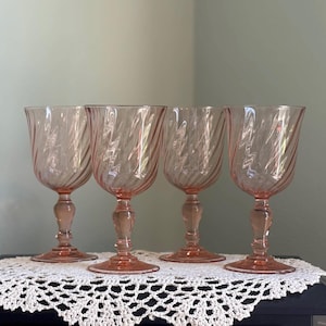 Pode incluir: Um conjunto de quatro taças de vidro rosa a combinar, cada uma com um padrão de espiral distinto na taça e um pé decorativo facetado. O vidro é translúcido com um tom rosa claro e uma base circular larga e estável.