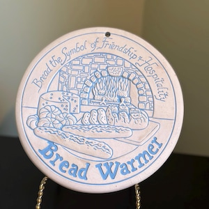 Peut inclure: Un chauffe-pain rond en céramique blanc cassé avec une illustration bleue en relief représentant une cheminée, divers pains et le texte "Bread the Symbol of Friendship & Hospitality" en haut et "Bread Warmer" en bas de l'objet.