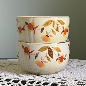 Puede incluir: Dos cuencos de cerámica apilados de color crema con un patrón floral repetitivo en tonos naranja, amarillo y marrón. Cada cuenco tiene una sutil textura acanalada vertical y un borde fino de tono dorado en la parte superior.