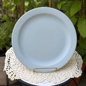 Puede incluir: Un plato redondo de cerámica azul claro con un acabado mate suave y un borde ancho y plano. El plato se muestra en posición vertical sobre un soporte de metal dorado fino, descansando sobre un tapete de encaje de ganchillo blanco.