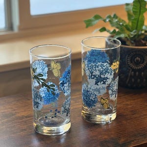 Puede incluir: Dos vasos transparentes con un diseño floral y de mariposas. Los vasos presentan flores de hortensia azules y mariposas amarillas. Los vasos son cilíndricos y se apoyan sobre una superficie de madera.