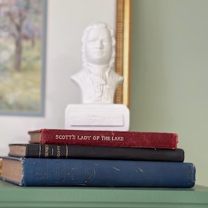 May include: A white bust of a man sits atop a stack of books. The books are in various colors, including red, black, and blue. The top book reads "SCOTT'S LADY OF THE LAKE". The bust is of a man with a powdered wig.