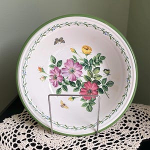May include: A white ceramic serving bowl with a green rim and floral design. The bowl features pink and yellow roses, green leaves, and butterflies. A green vine design with small blue flowers decorates the rim.