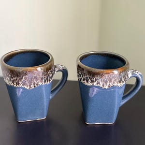 Puede incluir: Dos tazas de cerámica azules con forma cuadrada y borde marrón texturizado. Las tazas tienen un asa y un interior azul oscuro. Las tazas están colocadas una al lado de la otra.