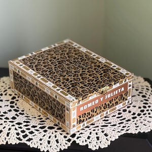 May include: Rectangular wooden box with a leopard print design on the lid and sides. The box features the text "ROMEO Y JULIETA" in a copper-colored font. The edges are trimmed with gold and white detailing.