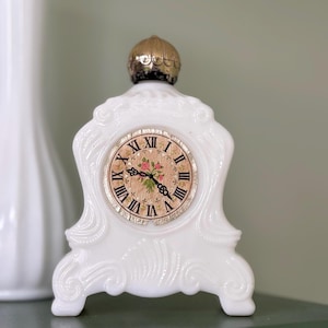 May include: A white, vintage-style mantel clock with a floral clock face. The clock has Roman numerals and a gold-colored top. The clock is made of a white, possibly ceramic material, and has ornate detailing.