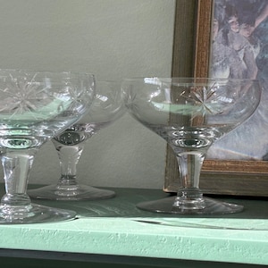 May include: A set of clear glass coupe glasses featuring a delicate starburst pattern etched into the bowl. Each glass has a wide, shallow bowl, a short stem, and a circular base, designed for serving beverages with a classic, transparent aesthetic.