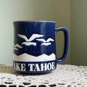 May include: A dark blue ceramic mug featuring a white embossed design of flying seagulls above a mountain range. The text LAKE TAHOE is printed in white capital letters along the bottom. The mug has a brown rim and a matching dark blue handle.