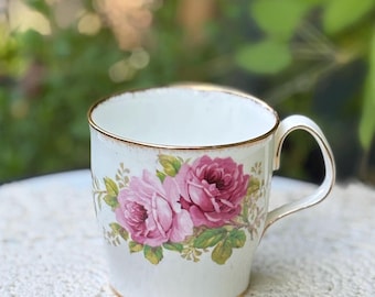 May include: A white ceramic mug featuring a floral pattern of pink and magenta roses with green leaves. The mug has a gold-coloured rim along the top edge and a matching gold-coloured trim on the base and the handle, providing a classic appearance.