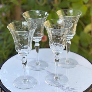 May include: A set of four clear glass stemmed goblets, each featuring a delicate etched floral and leaf pattern around the upper bowl. The glasses have slender stems and circular bases, showcasing a transparent design with fine detailing.