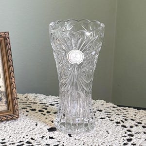 Puede incluir: Un jarrón de cristal tallado transparente con una parte superior acampanada y patrones geométricos intrincados. El jarrón tiene una base redonda y una etiqueta cerca del centro. El jarrón es probablemente para fines decorativos.