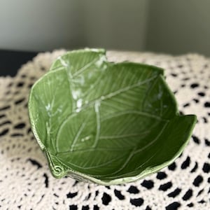 May include: A green ceramic leaf-shaped bowl with detailed vein patterns. The bowl has a glossy finish and is set against a neutral background. This decorative bowl is ideal for serving snacks or as a unique home decor accent.
