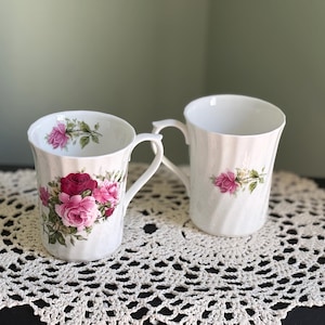 May include: Two white ceramic mugs with a floral rose design. One mug features a cluster of red and pink roses, while the other has a single pink rose. Both mugs have a ridged texture and curved handles.