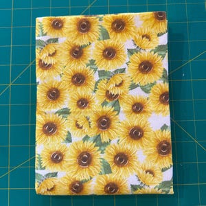May include: A rectangular piece of fabric featuring a repeating pattern of yellow sunflowers with brown centers and green leaves on a white background. The material is folded into a neat stack, displaying the floral print across the surface.