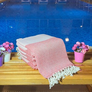 Peut inclure: Une pile de serviettes de plage à motifs roses et blancs avec des franges blanches. Les serviettes sont exposées sur un banc en bois à côté d'une piscine. De petits arrangements floraux dans des seaux violets, blancs et roses sont également sur le banc.