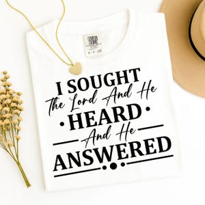 Puede incluir: Camiseta blanca con el texto "I Sought The Lord And He Heard And He Answered" en escritura negra. Se ven un collar con colgante de corazón dorado y un sombrero beige. La camiseta tiene una etiqueta que dice "COCOON COUSINS S-P-CH".
