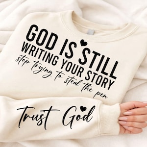 以下が含まれることがあります： クリーム色のスウェットシャツ。黒字で「GOD IS STILL WRITING YOUR STORY stop trying to steal the pen」と書かれています。袖には筆記体で「Trust God」と書かれています。「still」と「God」の上には小さな黒いハートがあります。
