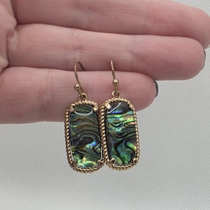 May include: A pair of gold-colored earrings with rectangular abalone shell inlays. The iridescent shells display shades of green, blue, and purple. The earrings have a textured, beaded border and a hook closure.