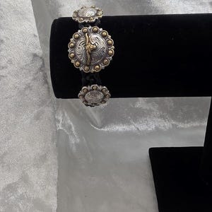 May include: Silver and gold-toned bracelet with three decorative circular medallions. The central medallion features a detailed design with a gold-toned figure. The bracelet is displayed on a black stand against a white background.