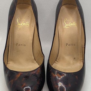 May include: A pair of high-heeled pumps featuring a glossy brown and black tortoiseshell pattern. The interior insoles are beige and display the gold-printed text Christian Louboutin Paris. The shoes have a rounded toe and a polished finish.