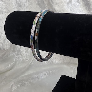 Puede incluir: Dos brazaletes finos de metal con patrones de esmalte de colores. Uno tiene una base blanca con motivos florales en azul, verde y naranja, mientras que el otro tiene una base azul oscuro con detalles decorativos de esmalte multicolor.